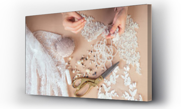 Wizualizacja Obrazu : #211729429 zbliżenie dłoni kobiety krawcowej ( krawcowa) projektantki sukni ślubnej przyszywającej koraliki do koronki na niebieskim tle w pracowni