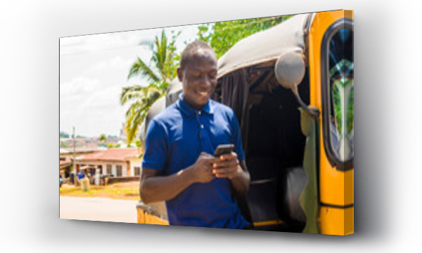wesoły afrykańczyk stojący obok swojej taksówki tuk tuk uśmiechający się i używający smartfona