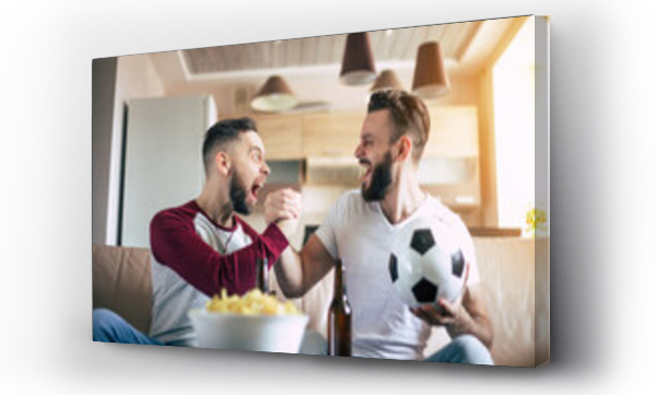 Wizualizacja Obrazu : #428784071 Dwóch najlepszych przyjaciół i fanów piłki nożnej oglądających jakiś mecz sportowy w telewizji, pijących piwo i jedzących przekąski podczas kibicowania drużynie na kanapie