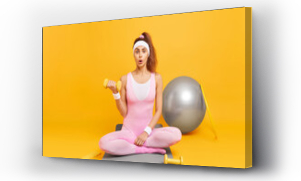 Wizualizacja Obrazu : #436341753 Full length shot of surprised woman has fitness practice lifts dumbbell poses crossed legs on karemat does exercises in sports club isolated over yellow background. People wellness and sport