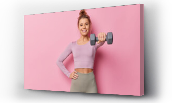 Wizualizacja Obrazu : #480701273 Sporty young woman being in good physical shape keeps hand on waist raises dumbbell has regular fitness training wears sportsclothes poses against pink background. Gym workout and sport concept