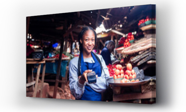 Wizualizacja Obrazu : #538883311 African millennial lady wearing colourful Apron selling tomatoes and vegetables in a typical local african market holding a POS terminal