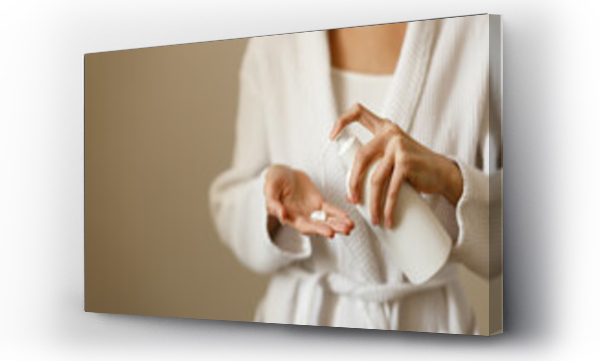 Wizualizacja Obrazu : #573270250 Young woman in white housecoat pours lotion or hand moisturizer from white bottle into the palm of her hand. Graceful hands of woman in close-up. Healthy body care after shower.