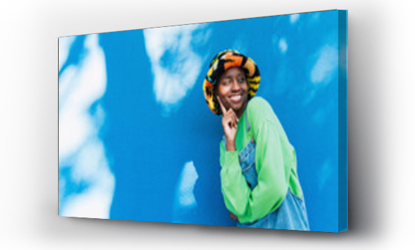 Wizualizacja Obrazu : #656967823 Happy young woman wearing colorful hat touching face in front of blue wall