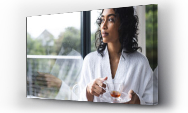 Wizualizacja Obrazu : #669029050 Happy biracial woman in bathrobe holding cup of tea and looking out window in sunny room at home